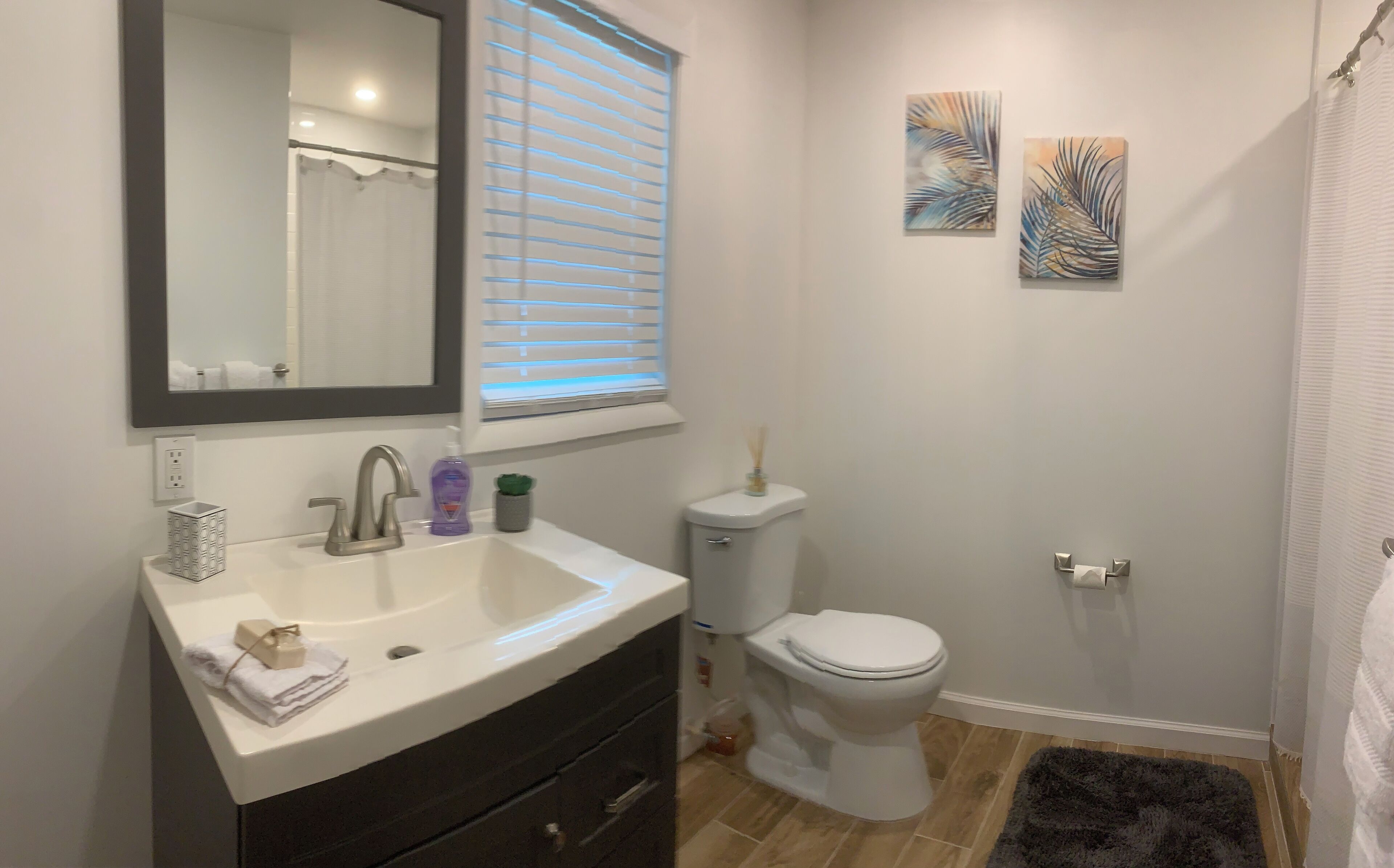 A bathroom with a white sink, dark vanity, a toilet, and a shower curtain. There are two wall-mounted paintings and a window with blinds.
