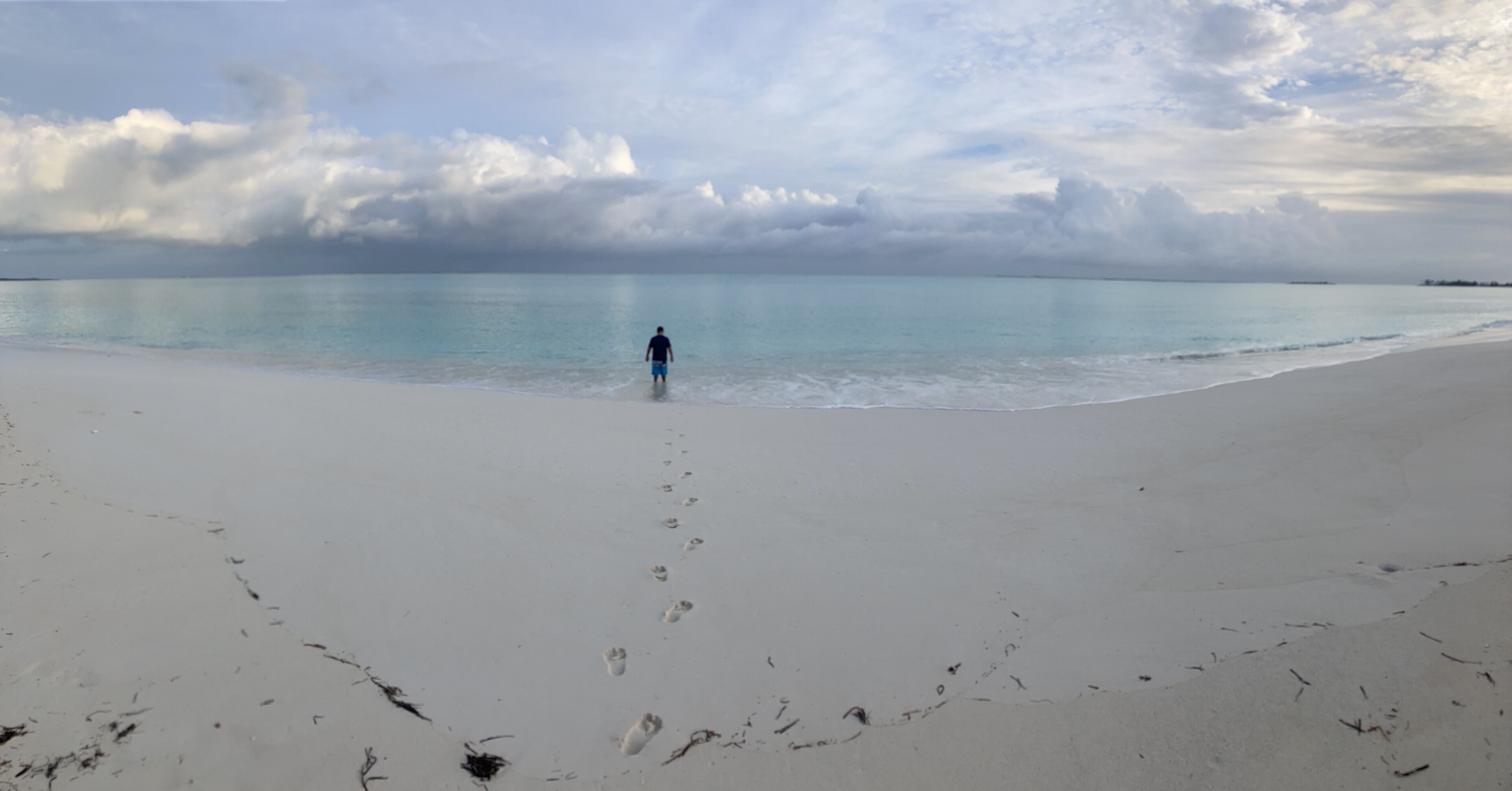 A beach with footprints leading to the water, a person standing in the distance, and a cloudy sky.