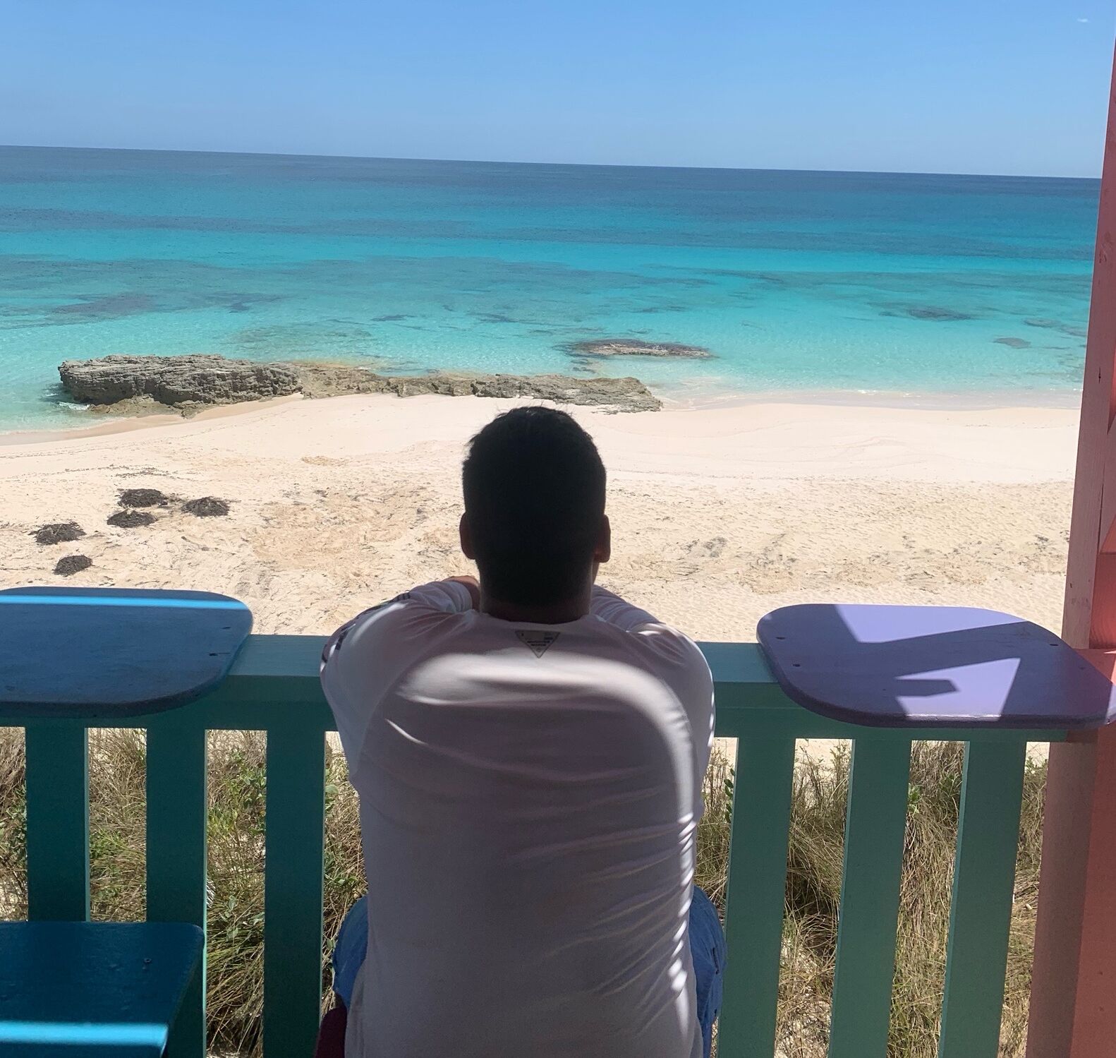 Una persona sentada en un banco admirando una playa con agua turquesa cristalina y una orilla arenosa.