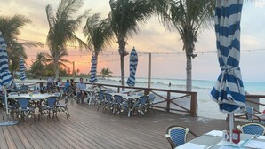 Een houten terras met tafels en stoelen, uitkijkend op een strand met palmbomen en een ondergaande zon.