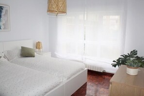 A bedroom with a bed, a bedside table, a lamp, a window with sheer curtains, and a potted plant.