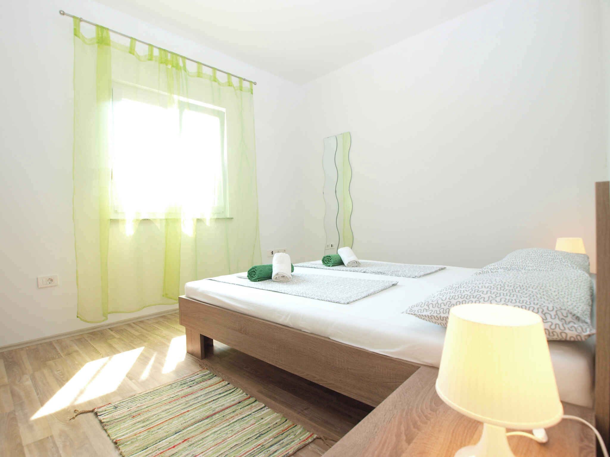 A bedroom with a bed, a bedside table with a lamp, a green towel, a white towel, and a window with sheer curtains.