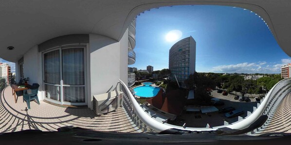 A balcony with a view of a swimming pool, a tall building, and a clear sky.