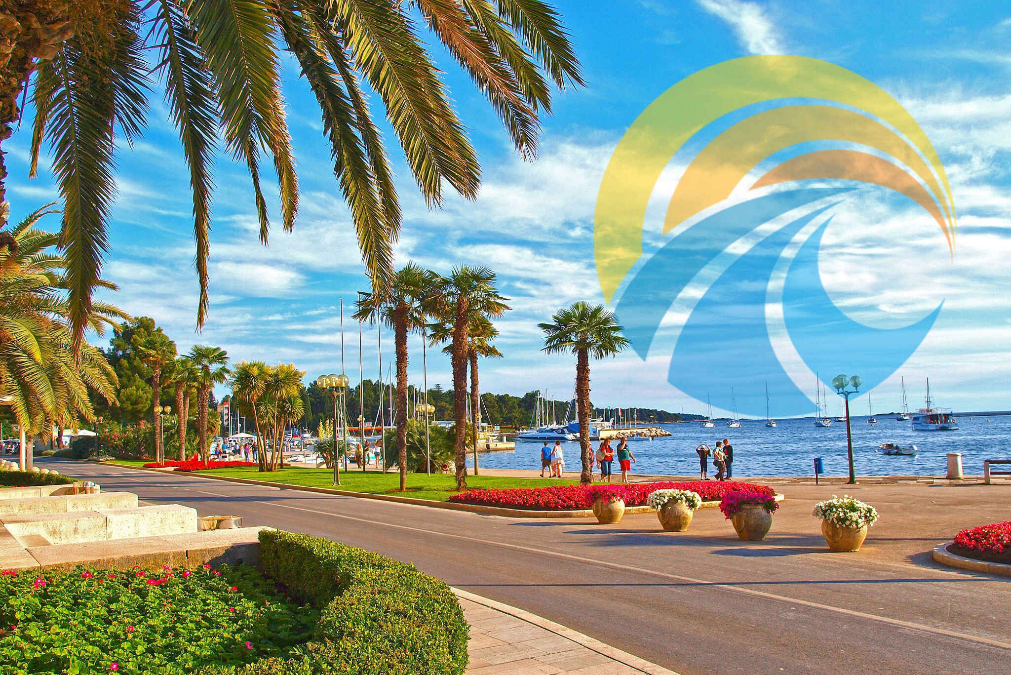 A coastal promenade with palm trees, flower beds, and a marina with boats.