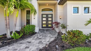 A modern house with a paved entrance, a double door, and a small garden with various plants. - Gulf Access, Putting Green, Outdoor Kitchen, Heated Pool & Spa & MORE! - Villa Captain's Quarters (Cape Coral)