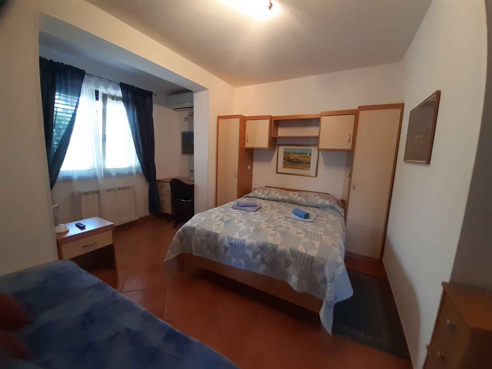 A bedroom with a bed, a desk, and a chair. There is a window with curtains, a radiator, and a picture on the wall.