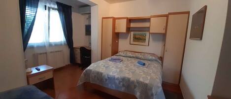 A bedroom with a bed, a desk, and a chair. There is a window with curtains, a radiator, and a picture on the wall.