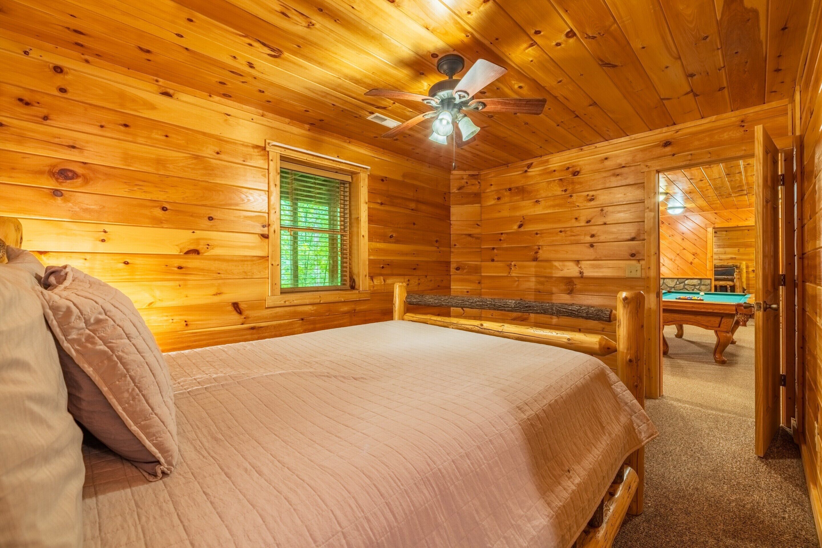 A wooden cabin room with a bed, a ceiling fan, a window with blinds, and a pool table in the background.