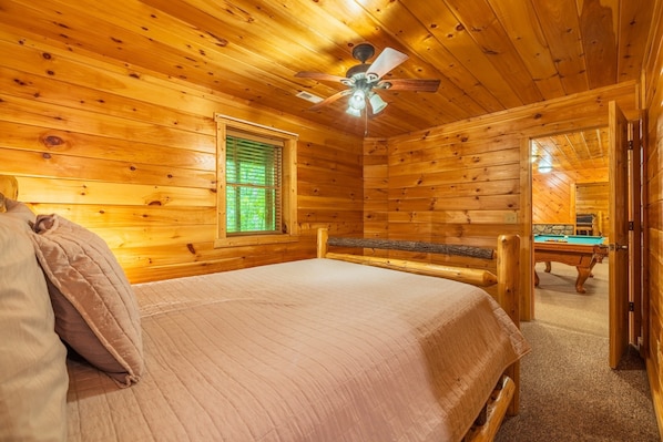 Habitación de cabaña de madera con una cama, un ventilador de techo, una ventana con persianas y una mesa de billar al fondo.