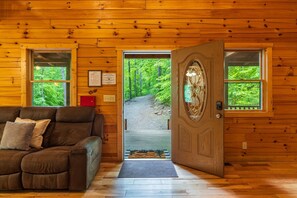 A wooden cabin interior with a brown leather sofa, a wooden door leading outside, and a window with a view of a forest path.