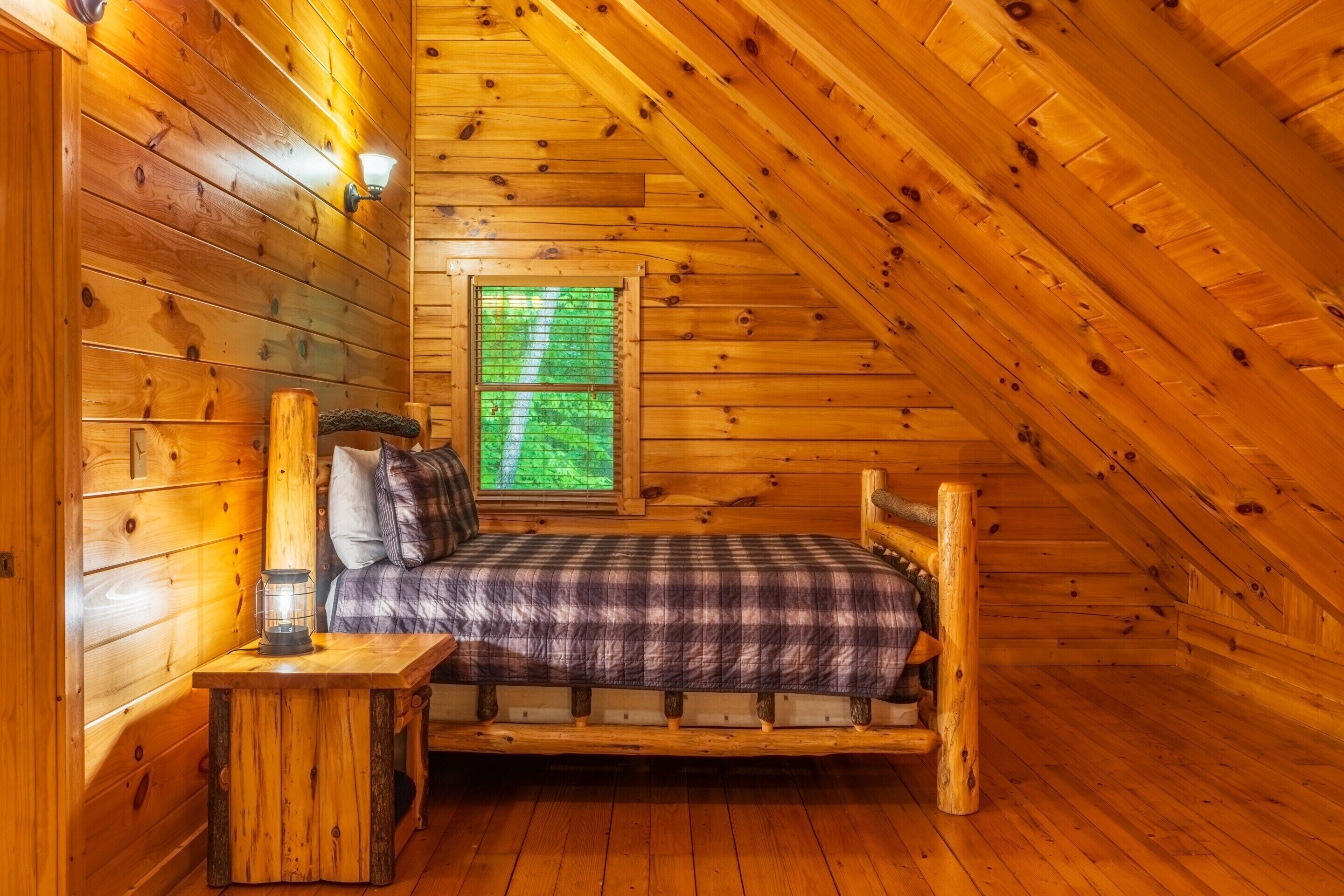 A cozy log cabin bedroom with a bed, a nightstand, a lantern, and a window with green foliage view.