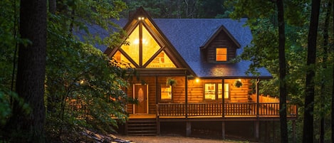 A log cabin with a porch, surrounded by trees.