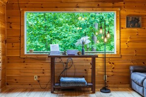 Interior de una cabaña de madera con un ventanal grande, un escritorio con una computadora portátil, una lámpara y un sofá.