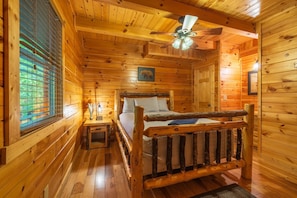 Camera da letto in una baita di legno, con un letto, un comodino, una ventola a soffitto e una finestra con persiane.