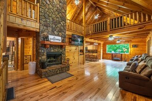 A log cabin interior with a stone fireplace, wooden floors, a balcony, and a television.