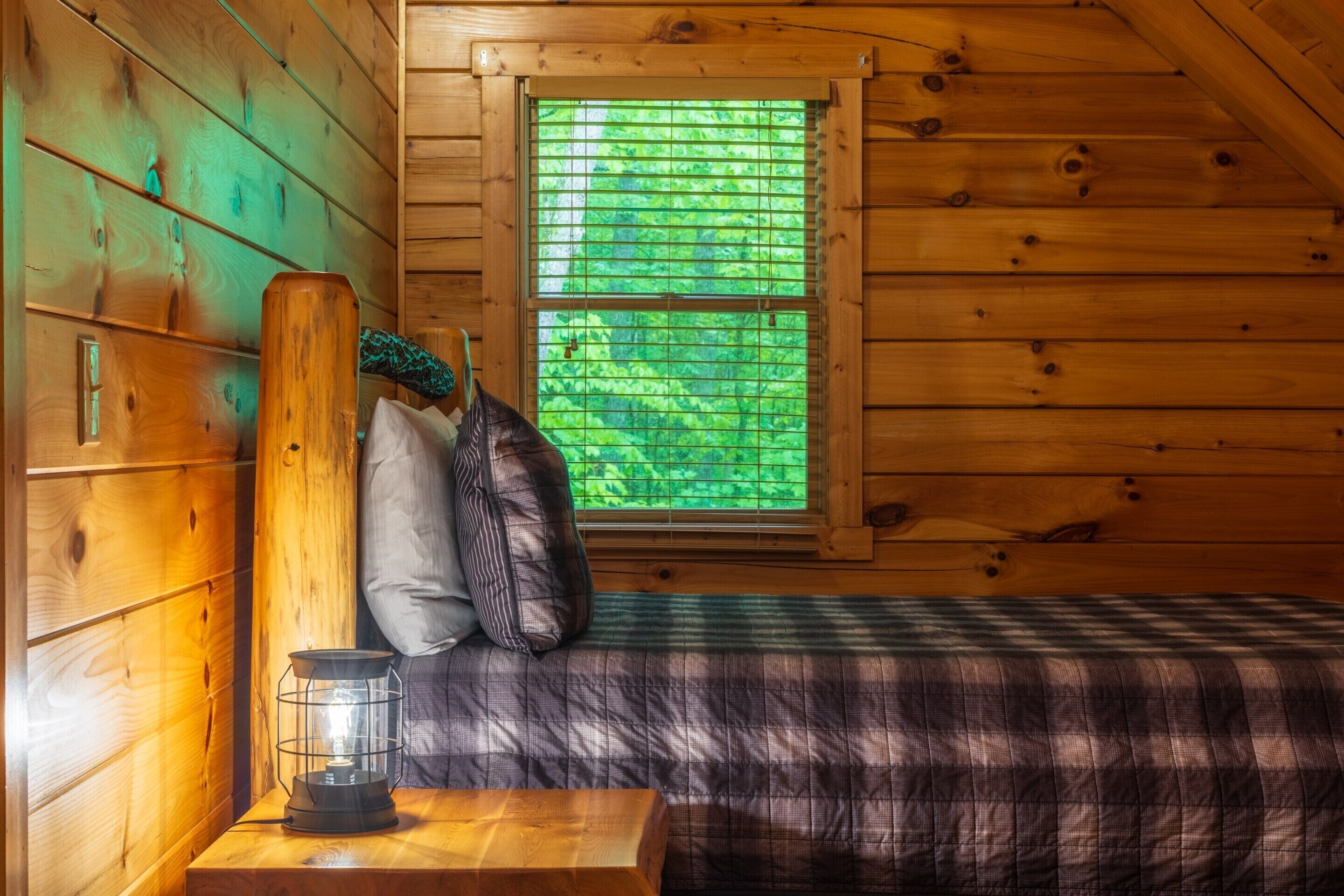 A wooden cabin room with a bed, pillows, a table with a lantern, and a window with blinds.