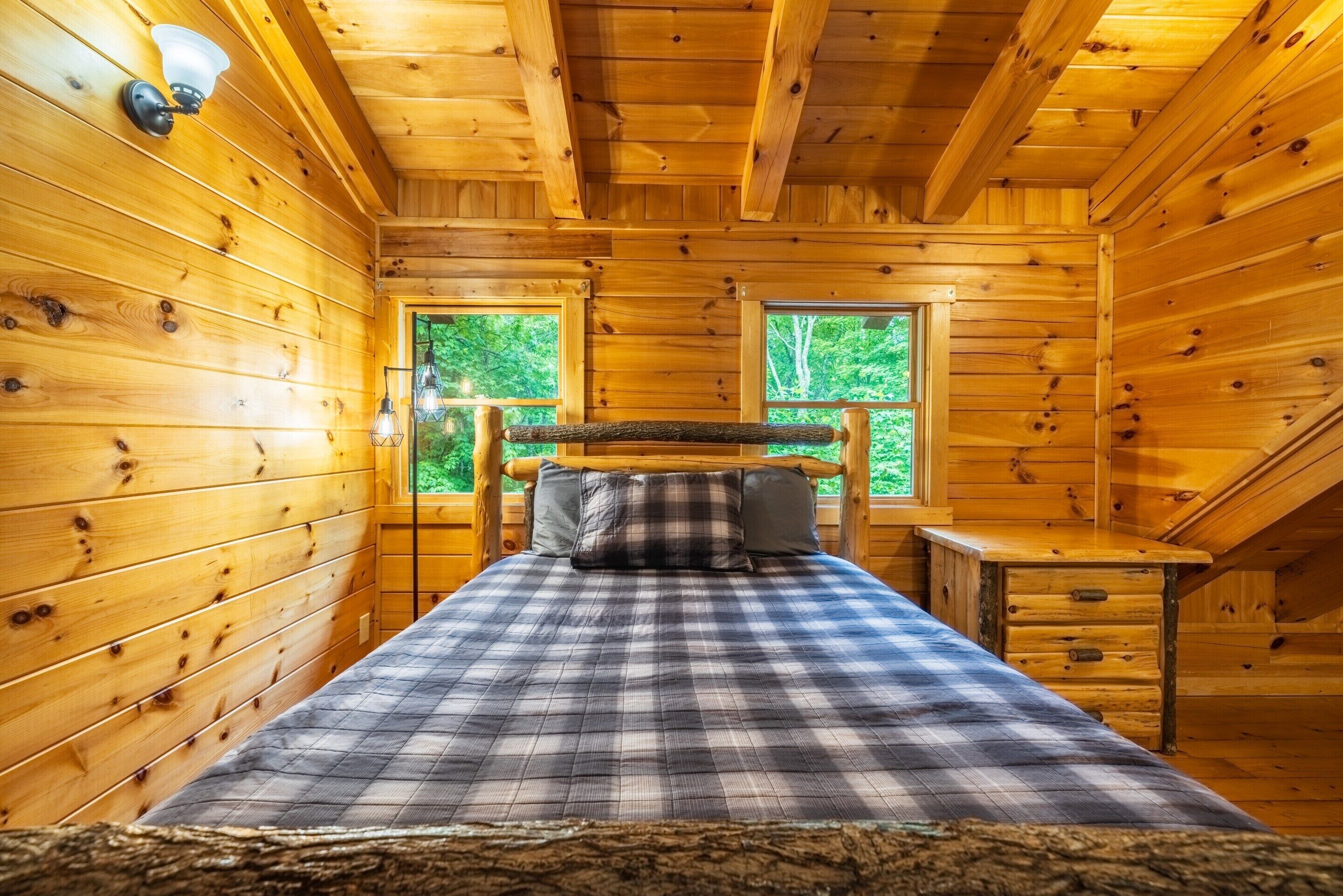 Habitació d'una cabana de fusta amb un llit, dues finestres i un escriptori petit.