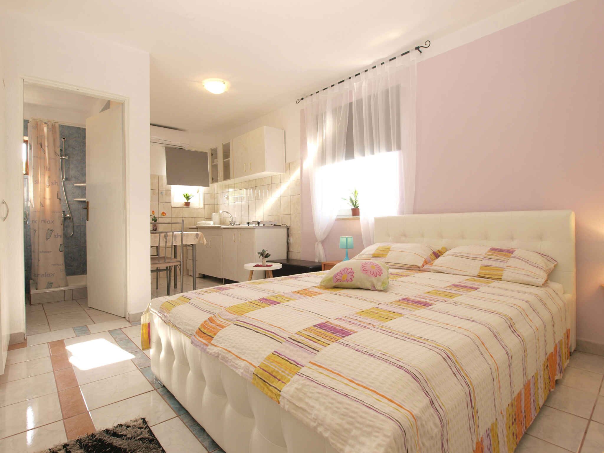 Een slaapkamer met een groot bed, een klein tafeltje, een raam met gordijnen en een kitchenette op de achtergrond.
