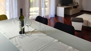 A dining area with a table set for two, a bottle of wine, and two wine glasses. There is a television in the background, a sofa, and a clock on the wall.