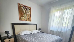 A bedroom with a bed, a nightstand, a drawer, a painting, and a window with curtains.