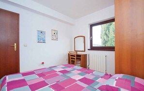 Quarto com cama, cadeira de madeira, cômoda, espelho e janela com vista para árvores.