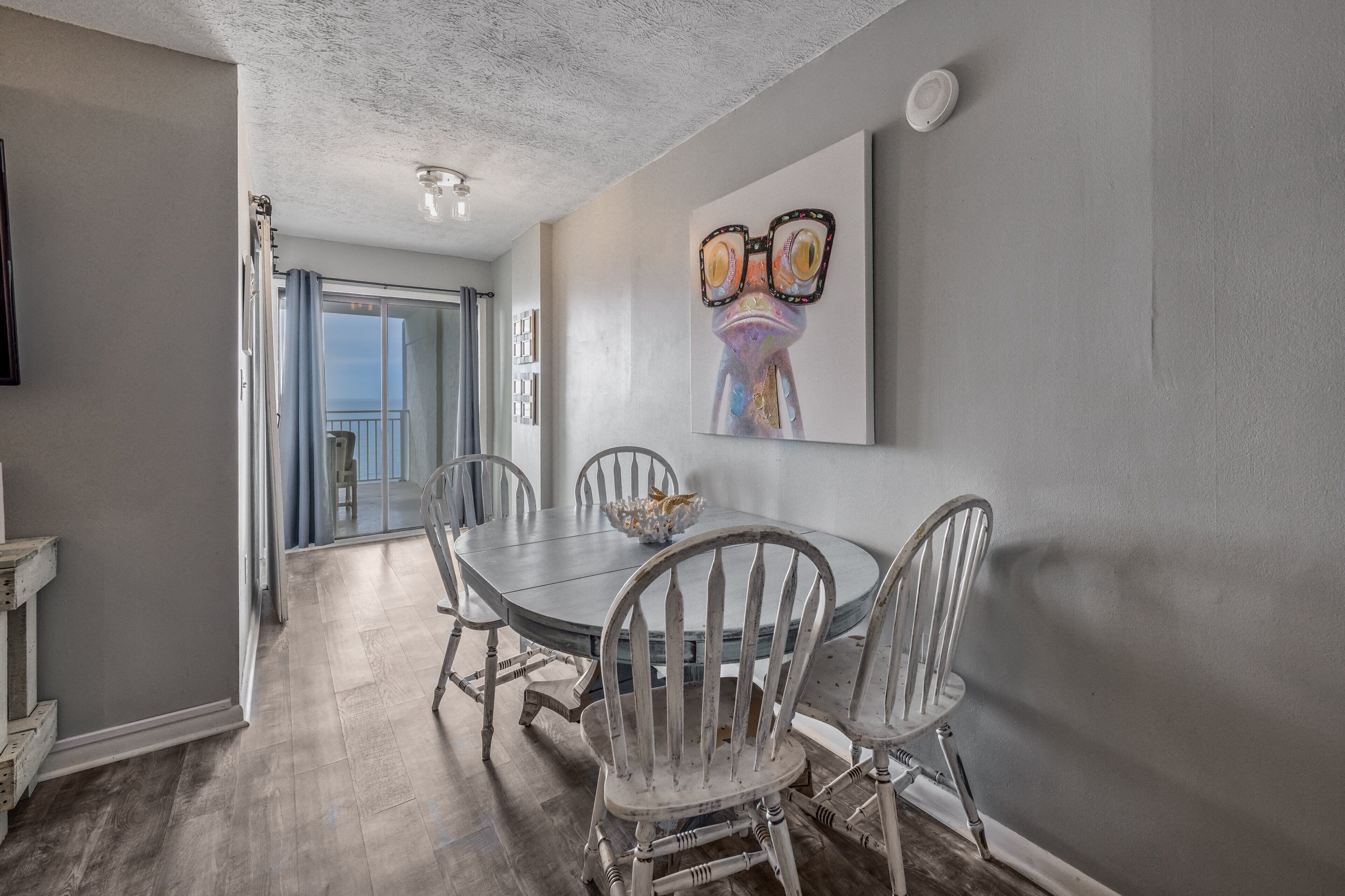 Un coin repas avec une table ronde, des chaises blanches et un tableau représentant une grenouille accroché au mur.