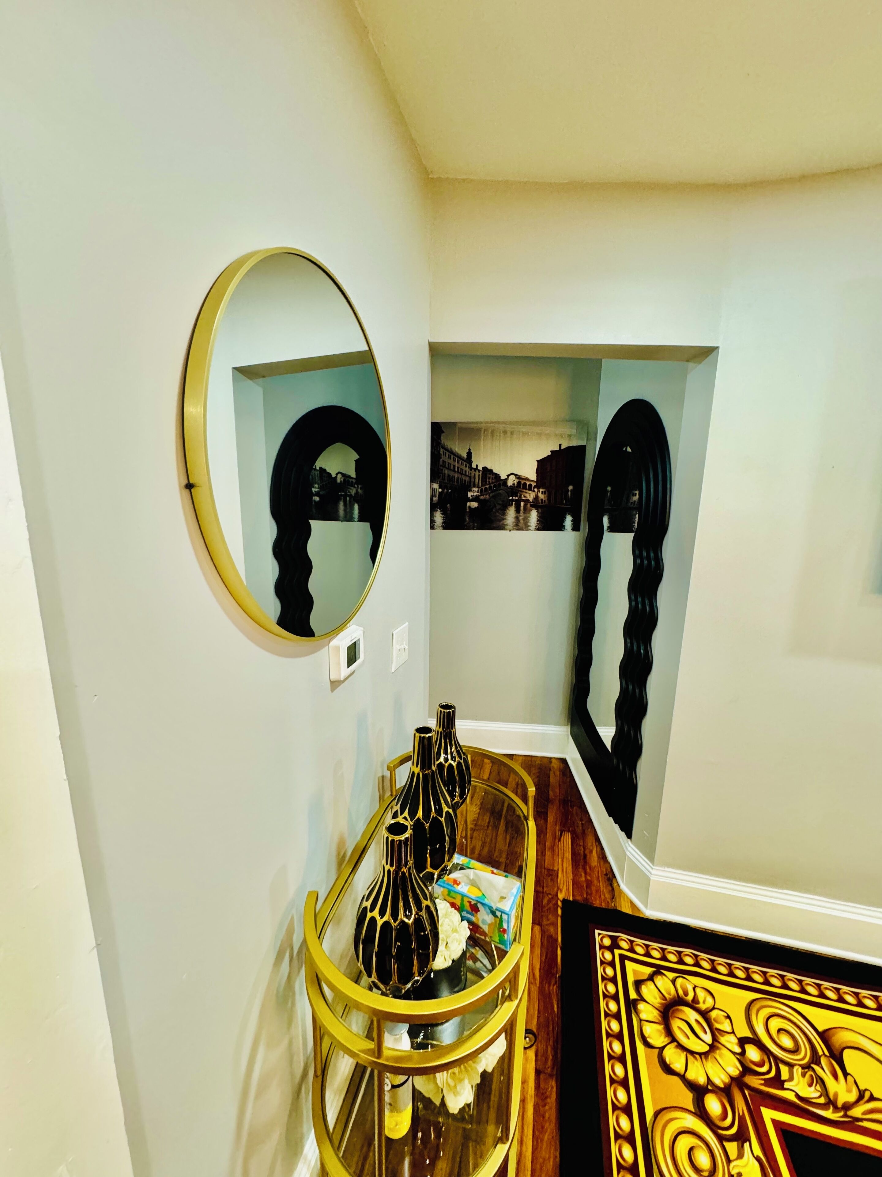 Un pasillo con un espejo redondo, una fotografía enmarcada y un jarrón decorativo sobre un soporte dorado.