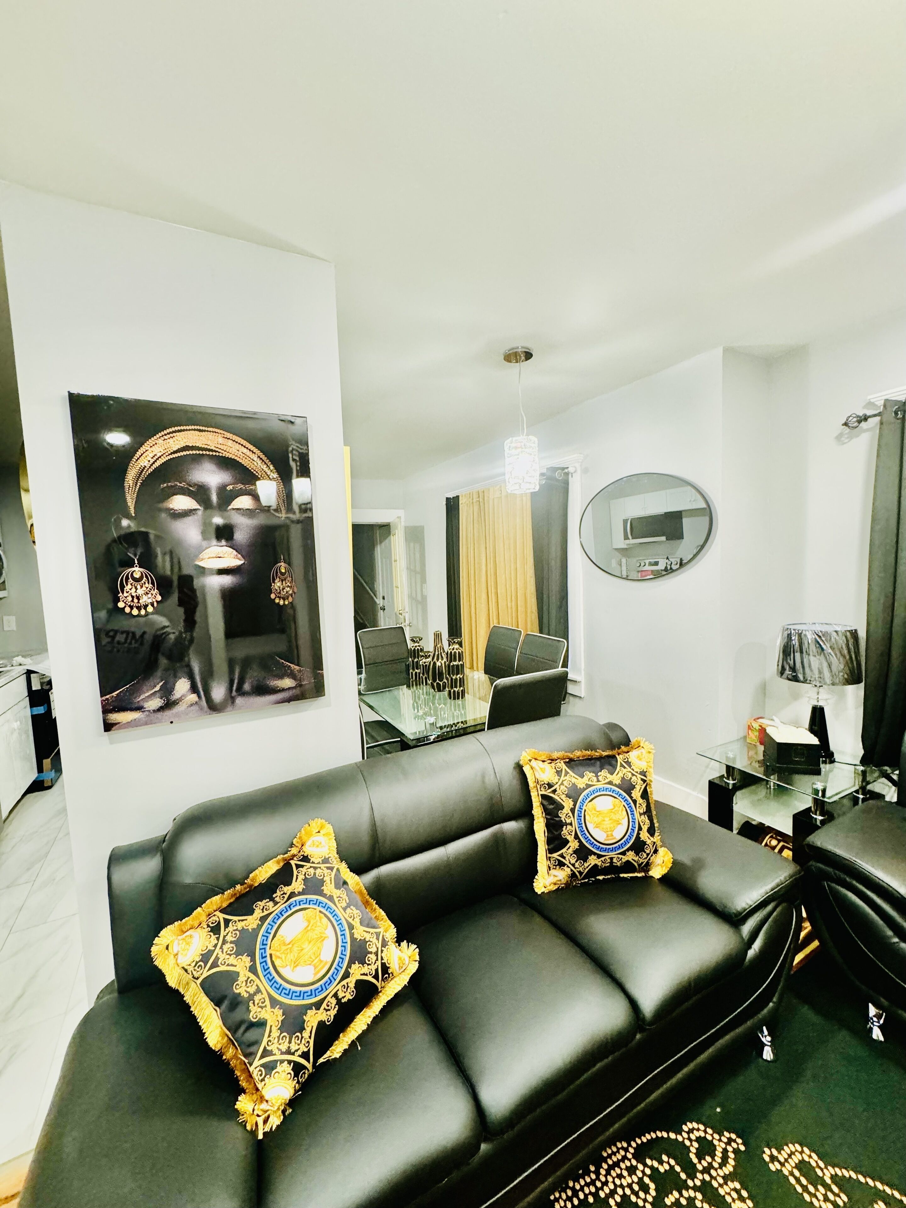 Un soggiorno moderno con un divano in pelle nera, due cuscini decorativi, un tavolino in vetro, uno specchio rotondo e, sullo sfondo, una zona pranzo.