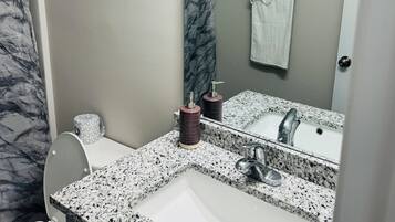 Un bagno con un lavandino bianco, un piano di lavoro in granito e uno specchio sopra. Si vede un WC con tenda da doccia a effetto marmo e un asciugamano su un porta asciugamani.