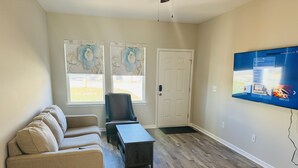 A living room with a beige sofa, a black armchair, and a coffee table. There is a door and windows with decorative floral panels. A flat-screen TV is mounted on the wall. - Brand NEW Entire home in Statesville, NC (Statesville)