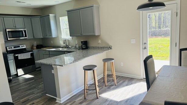 A modern kitchen with grey cabinets, a granite countertop, and a dining area with a table and bar stools. - Brand NEW Entire home in Statesville, NC (Statesville)
