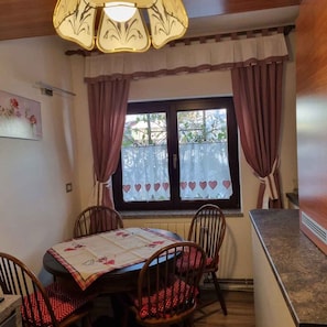 Uma sala de jantar com uma mesa posta para quatro pessoas, cadeiras de madeira, um candelabro e uma janela com cortinas.