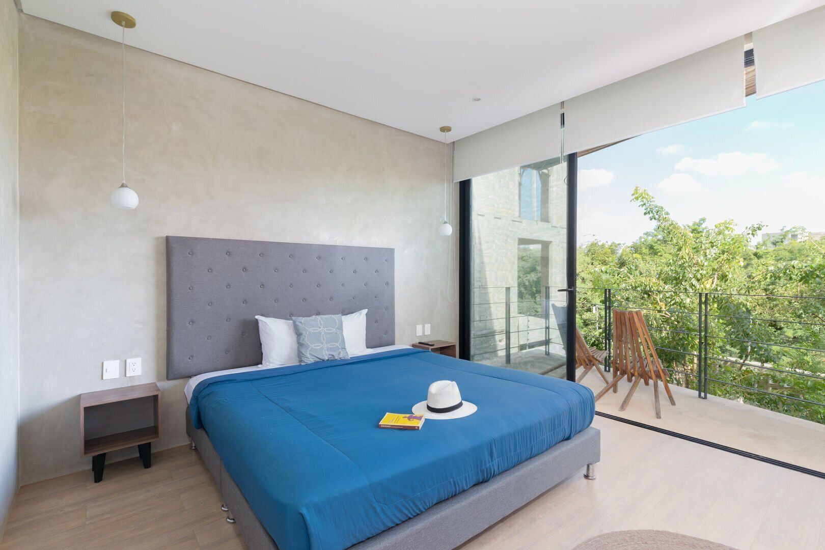 A modern bedroom with a large bed, a grey headboard, and a view of greenery through large windows.