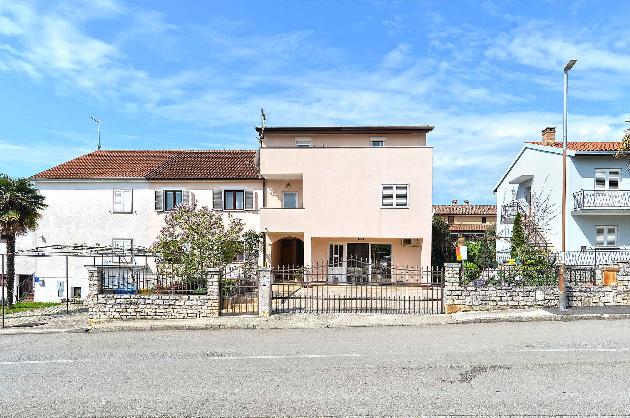 A two-story building with a stone wall and metal gate, featuring a flat roof and a balcony on the second floor.