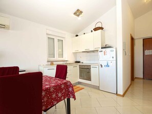 Una cocina compacta con armarios blancos, horno integrado y refrigerador. Hay una mesa de comedor con mantel rojo y dos sillas.