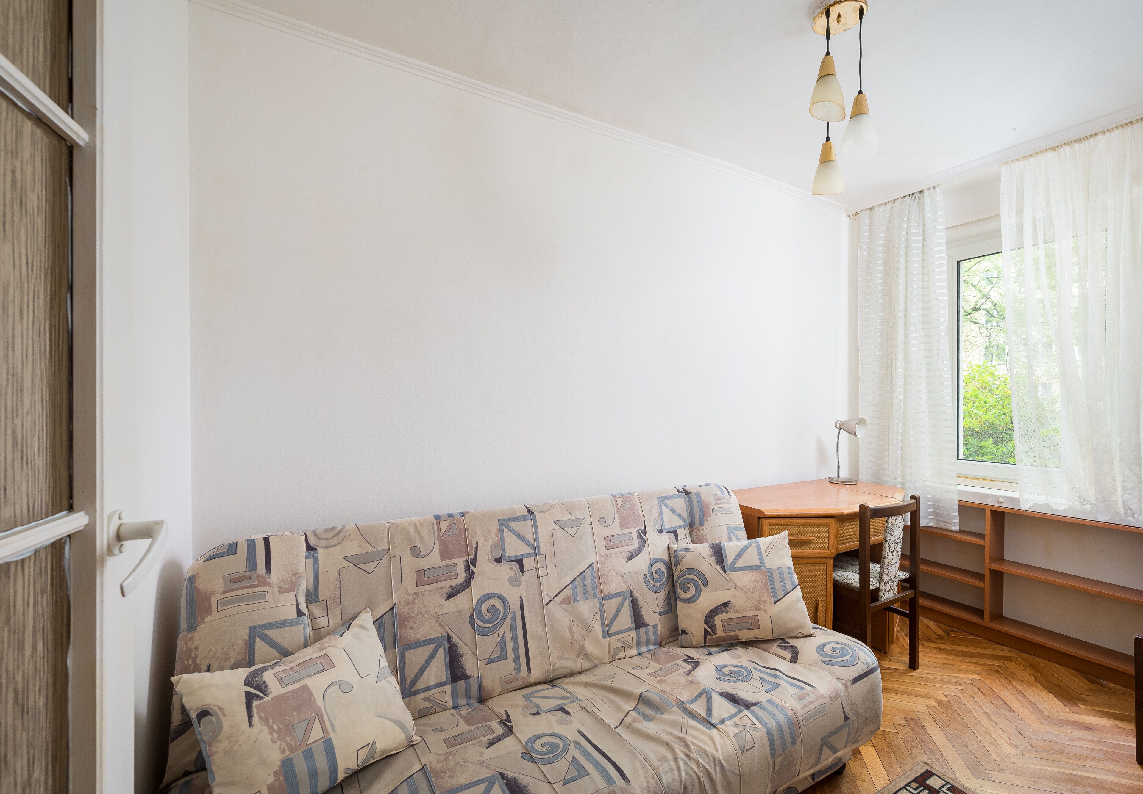 A living room with a beige sofa, patterned cushions, a wooden table, and a window with sheer curtains.