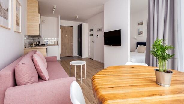 A modern apartment with a pink sofa, a wooden dining table, a small white stool, a potted plant, and a TV mounted on the wall. - Nova Letnica Apartment (Gdansk)