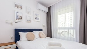 A neatly made bed with white linens and beige pillows, a bedside table with a lamp, and a window with a view of buildings. - Nova Letnica Apartment (Gdansk)