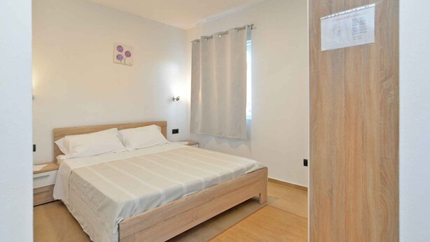 A bedroom with a wooden bed, a nightstand, a wall-mounted light, a window with curtains, and a door with a sign.