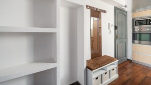 Un pasillo con paredes blancas, un banco de madera y una puerta que da a una zona de cocina con microondas y horno.