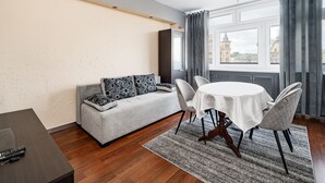 Una habitación de hotel moderna con un sofá gris, una mesa de comedor redonda y vistas de la ciudad a través de amplias ventanas.