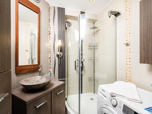 Un baño moderno con ducha de vidrio, tocador de madera con lavabo y lavadora Samsung.