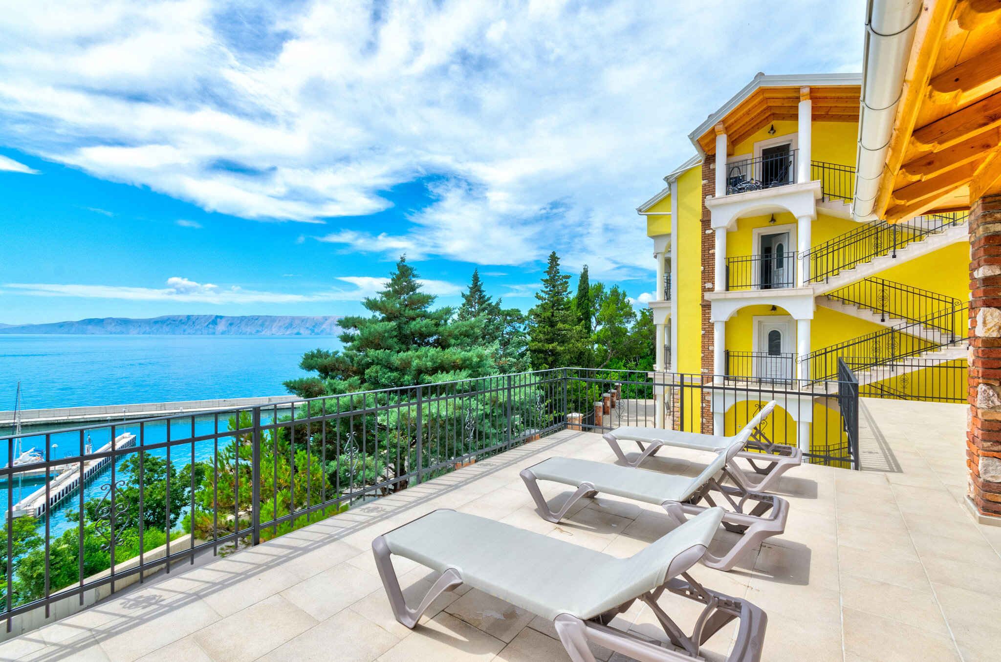 Un balcón con sillones de descanso que ofrece vistas al mar, un edificio amarillo con una escalera y un cielo despejado.