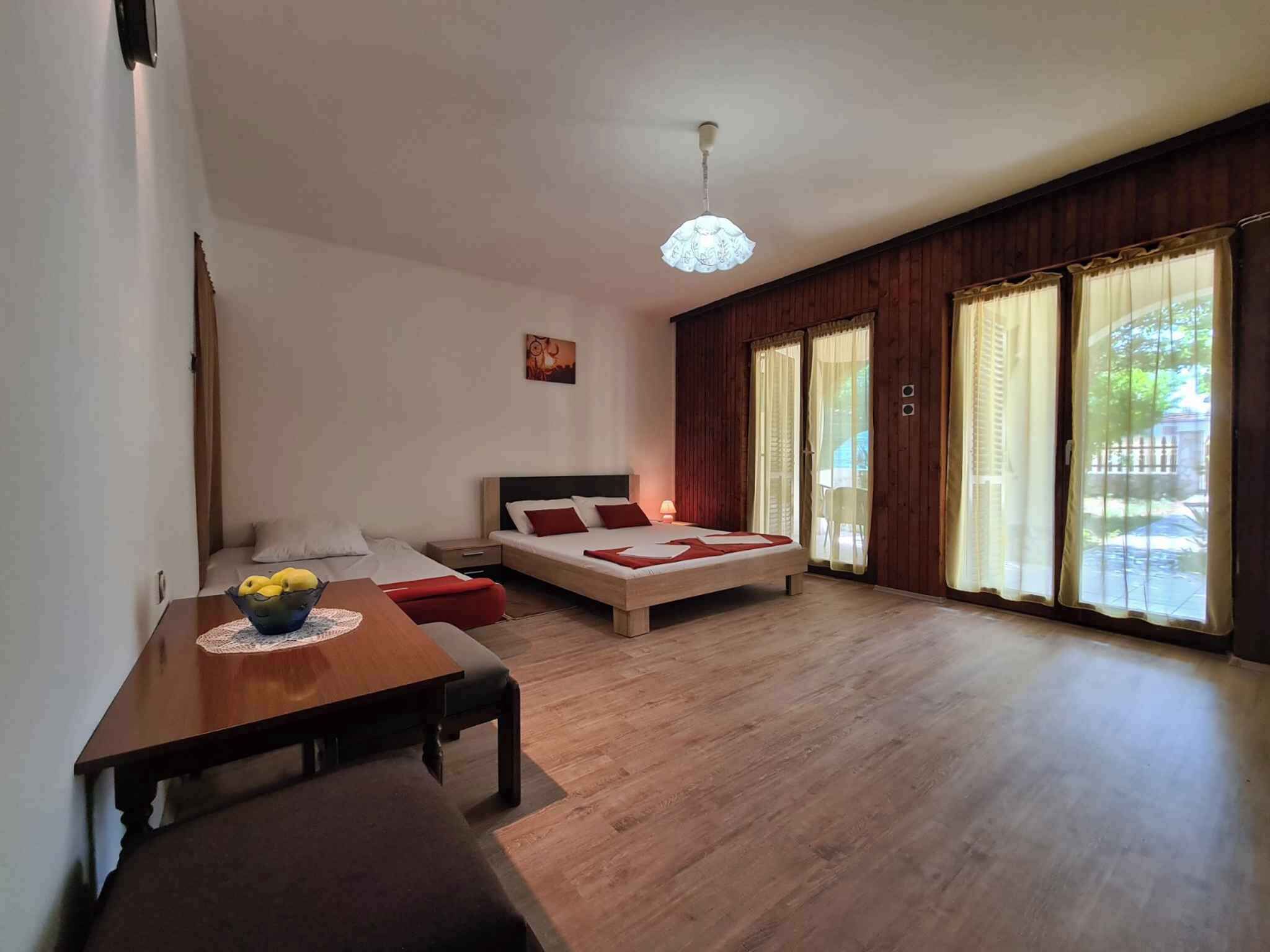 Una habitación amplia con piso de madera, una cama con ropa de cama roja, una mesa de comedor con un tazón de fruta y ventanas grandes con cortinas.