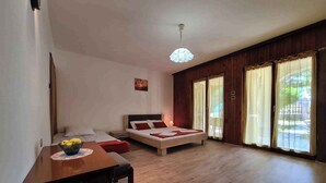 A spacious room with a wooden floor, a bed with red bedding, a dining table with a bowl of fruit, and large windows with curtains. - Holiday apartment mit Terrasse (Valbandon)