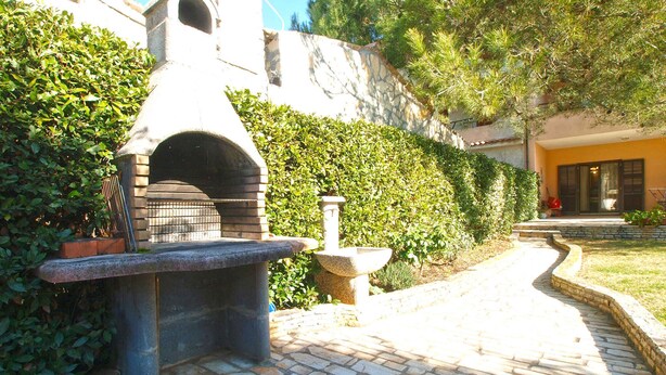 A stone outdoor kitchen with a chimney, surrounded by greenery and a paved pathway leading to a building. - Holiday apartment with air conditioning (Fažana)