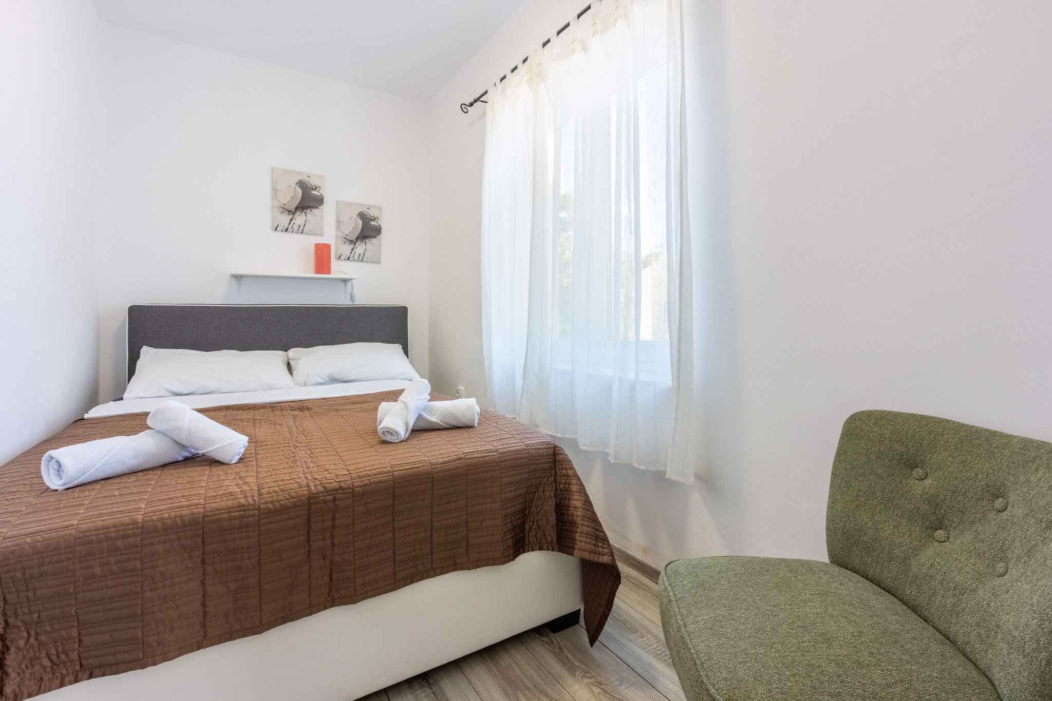 Ein ordentlich bezogenes Bett mit brauner Tagesdecke und weißen Kissen, ein grüner Sessel und ein Fenster mit durchsichtigen Vorhängen.