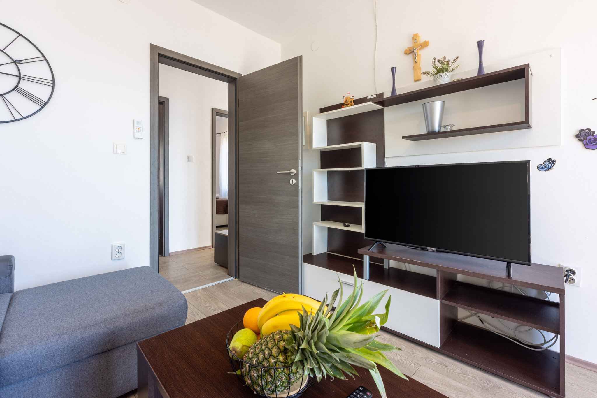 A modern living room with a grey sofa, a wooden coffee table with fruit, a flat-screen TV, and built-in shelves with decorative items.