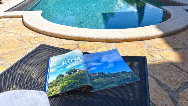 Une piscine bordée d’une terrasse en pierre, un magazine posé sur un tapis et des chaises longues.
