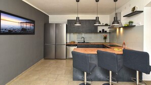 Une cuisine moderne dotée d’un grand réfrigérateur, d’un îlot central en bois avec des tabourets de bar, d’un éclairage suspendu et d’une vue sur la mer par une fenêtre.